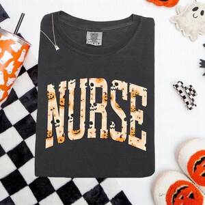 May include: A charcoal gray crewneck t-shirt featuring the word NURSE in large, arched block letters. The letters are filled with a repeating pattern of orange jack-o'-lanterns, skulls, and small stars against a light beige background.