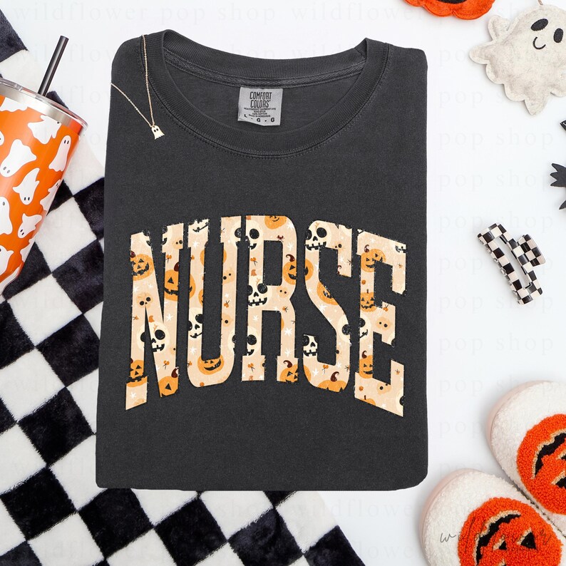 May include: A charcoal gray crewneck t-shirt featuring the word NURSE in large, arched block letters. The letters are filled with a repeating pattern of orange jack-o'-lanterns, skulls, and small stars against a light beige background.