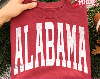 Puede incluir: Una camiseta de cuello redondo color granate con la palabra ALABAMA impresa en letras mayúsculas blancas grandes y desgastadas en el pecho. El diseño tiene una textura vintage y la camiseta está hecha de tela lisa con cuello acanalado.