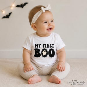 May include: A white short-sleeved t-shirt featuring the black text MY FIRST BOO with a small ghost graphic replacing the letter O. The shirt is paired with light beige ribbed pants and a matching white fabric headband with a small bow.