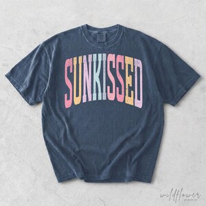 May include: A dark blue short-sleeve t-shirt featuring the word SUNKISSED printed in a distressed, arched font across the chest. The letters are colored in alternating shades of pink, yellow, light blue, and orange, creating a vibrant contrast.