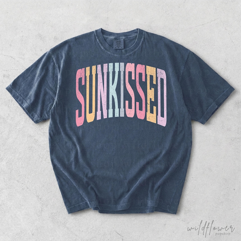 May include: A dark blue short-sleeve t-shirt featuring the word SUNKISSED printed in a distressed, arched font across the chest. The letters are colored in alternating shades of pink, yellow, light blue, and orange, creating a vibrant contrast.