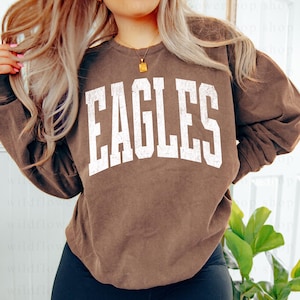 May include: A brown sweatshirt with the word "EAGLES" printed in large, distressed white letters across the chest. The sweatshirt has long sleeves and a crew neck. The overall style is casual and comfortable.