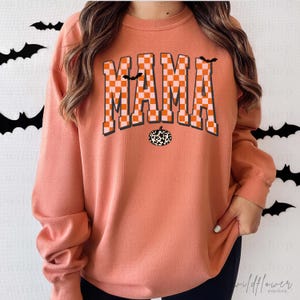 Puede incluir: Una sudadera de cuello redondo color melocot&oacute;n con la palabra MAMA en un patr&oacute;n de cuadros naranja y blanco. El dise&ntilde;o incluye dos peque&ntilde;os murci&eacute;lagos negros y una calabaza con estampado de vaca en blanco y negro bajo el texto.