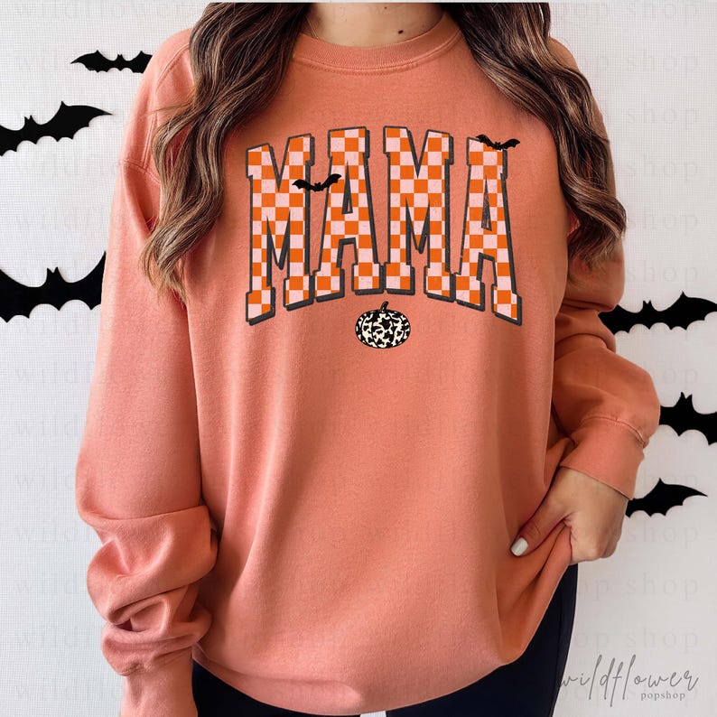 Puede incluir: Una sudadera de cuello redondo color melocot&oacute;n con la palabra MAMA en un patr&oacute;n de cuadros naranja y blanco. El dise&ntilde;o incluye dos peque&ntilde;os murci&eacute;lagos negros y una calabaza con estampado de vaca en blanco y negro bajo el texto.