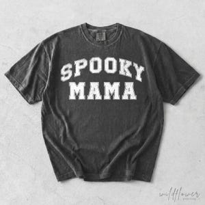 May include: A charcoal grey short-sleeve t-shirt with a distressed white collegiate-style graphic print on the chest. The text reads SPOOKY MAMA in a bold, arched font. The garment features a crew neckline and a vintage-inspired washed finish.