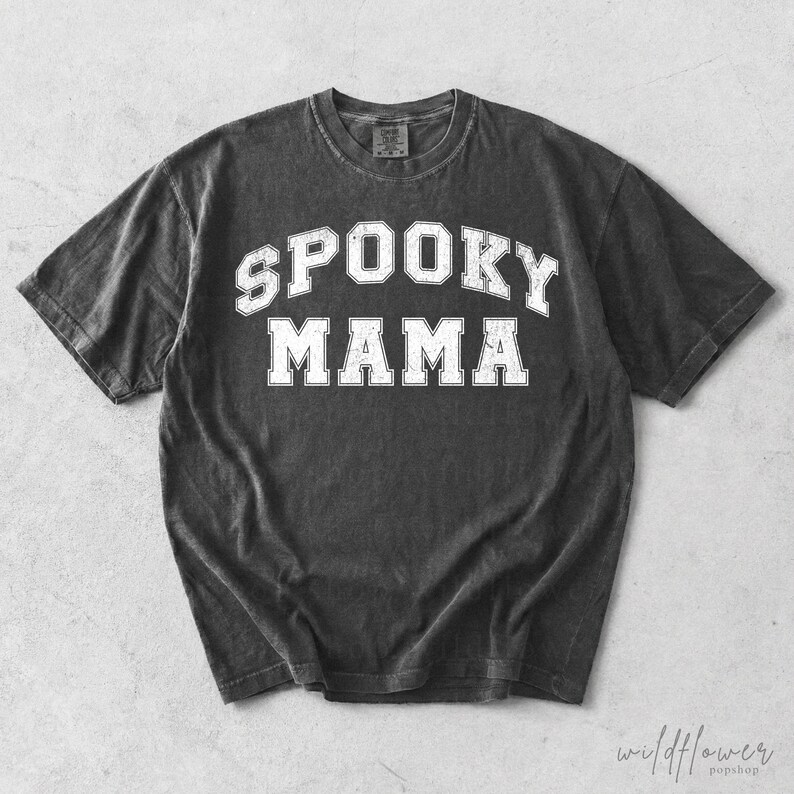 May include: A charcoal grey short-sleeve t-shirt with a distressed white collegiate-style graphic print on the chest. The text reads SPOOKY MAMA in a bold, arched font. The garment features a crew neckline and a vintage-inspired washed finish.