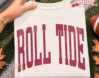 May include: A cream-colored crewneck sweatshirt featuring the text ROLL TIDE in a distressed, dark red collegiate font arched across the chest. The garment has a ribbed neckline and long sleeves, showcasing a clean, simple design on a neutral fabric.