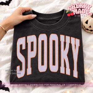 May include: A black crewneck sweatshirt featuring the word SPOOKY printed in large, arched, white and purple vertical striped letters with a distressed orange outline. The design is centered on the front of the garment, which has a faded, vintage-style texture.