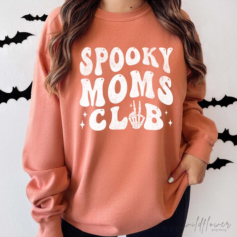 May include: A terracotta orange long-sleeve crewneck sweatshirt featuring white text that reads SPOOKY MOMS CLUB, with the letter L replaced by a white skeleton hand making a peace sign, accented by small white four-pointed star graphics.