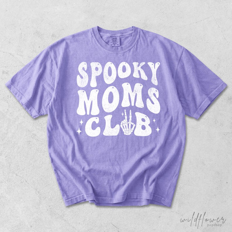 May include: A light purple short-sleeve t-shirt featuring the white distressed text SPOOKY MOMS CLUB printed on the front. The letter U in CLUB is replaced by a white graphic of a skeleton hand making a peace sign, surrounded by small stars.