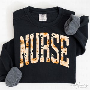 May include: A black long-sleeve sweatshirt featuring the word NURSE in large, block letters across the chest. The letters have a cream-colored background filled with a repeating pattern of orange jack-o'-lanterns and white skulls.