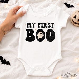 May include: A white short-sleeved baby bodysuit featuring the black text "MY FIRST BOO" printed on the chest. The letter "O" in "BOO" is replaced by a small, cream-colored ghost illustration. The design includes small gold star accents.
