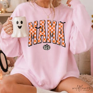Puede incluir: Una sudadera de manga larga rosa claro con la palabra MAMA en un patr&oacute;n de cuadros naranja y blanco. El dise&ntilde;o incluye peque&ntilde;as siluetas de murci&eacute;lagos negros y un gr&aacute;fico de calabaza con estampado de vaca en blanco y negro.
