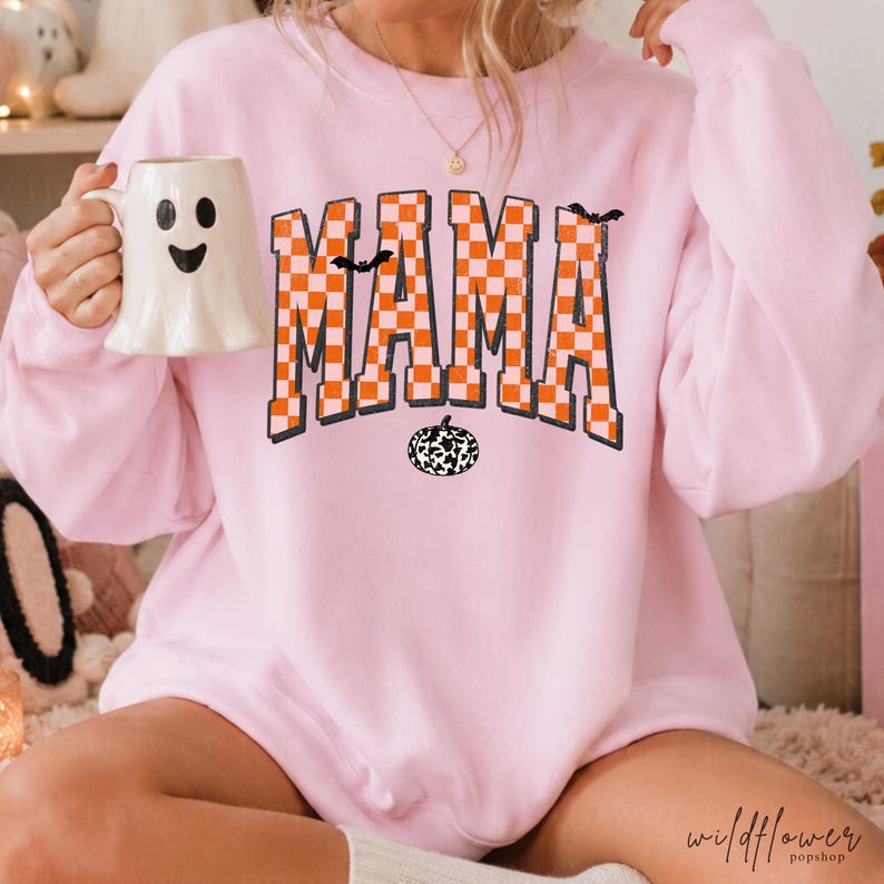 Puede incluir: Una sudadera de manga larga rosa claro con la palabra MAMA en un patr&oacute;n de cuadros naranja y blanco. El dise&ntilde;o incluye peque&ntilde;as siluetas de murci&eacute;lagos negros y un gr&aacute;fico de calabaza con estampado de vaca en blanco y negro.