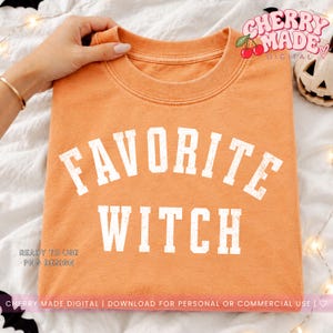 Op de afbeelding: Een oranje t-shirt met ronde hals en de witte, verweerde tekst "FAVORITE WITCH" in een vet, gebogen universiteitslettertype op de borst gedrukt. Het kledingstuk heeft een klassieke geribbelde halslijn en een zachte, textuurrijke stof.