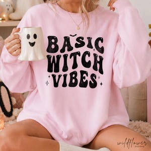 Pode incluir: Uma camisola de gola redonda rosa claro com texto preto que diz BASIC WITCH VIBES numa fonte ondulada, acentuada por um pequeno morcego e estrelas. A camisola é combinada com uma caneca de cerâmica branca em forma de fantasma.