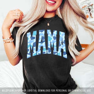 May include: A black short-sleeved t-shirt featuring the word MAMA printed in a large, arched, collegiate-style font. The letters are filled with a blue and light blue camouflage pattern. The shirt has a crew neckline and a relaxed fit.