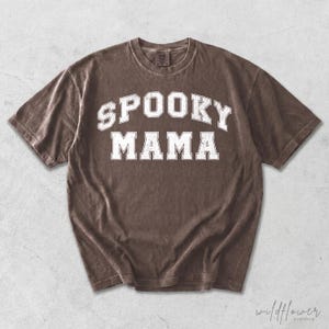 May include: A brown short-sleeve t-shirt featuring the text SPOOKY MAMA in a white, distressed, collegiate-style font across the chest. The garment has a crew neckline and a relaxed fit, with a visible tag at the inner collar area.