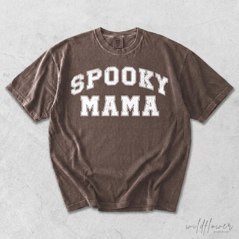 May include: A brown short-sleeve t-shirt featuring the text SPOOKY MAMA in a white, distressed, collegiate-style font across the chest. The garment has a crew neckline and a relaxed fit, with a visible tag at the inner collar area.