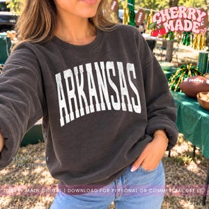 May include: A charcoal grey long-sleeve crewneck sweatshirt featuring the word ARKANSAS printed in a large, distressed white font across the chest. The garment has a ribbed neckline, cuffs, and hem, with a relaxed fit and a soft, worn-in texture.