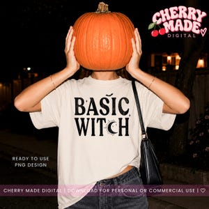 May include: A cream-colored t-shirt featuring the black text BASIC WITCH with a small bat, stars, and a crescent moon graphic. The shirt is worn with dark denim jeans and a black shoulder bag, while a large orange pumpkin obscures the face.