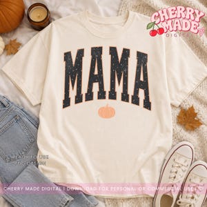 K&ouml;nnte beinhalten: Ein cremefarbenes T-Shirt mit dem Wort MAMA in gro&szlig;en, dunklen, abgenutzten Blockbuchstaben mit einem dezenten Sternenmuster. Unter dem Text befindet sich eine kleine, orangefarbene K&uuml;rbisgrafik. Das Shirt enth&auml;lt den Text CHERRY MADE DIGITAL.
