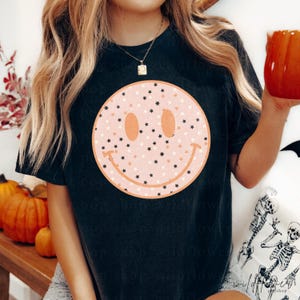 May include: A black short-sleeve t-shirt featuring a large, centered graphic of a light pink smiley face. The smiley face is filled with a pattern of small black and white stars and is outlined with a thick, solid peach-colored circular border.