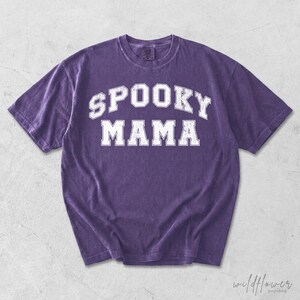 May include: A purple short-sleeve t-shirt with a crew neckline. The front features the text SPOOKY MAMA printed in a white, distressed, collegiate-style font. The fabric has a washed, vintage appearance with short sleeves and a standard hem.