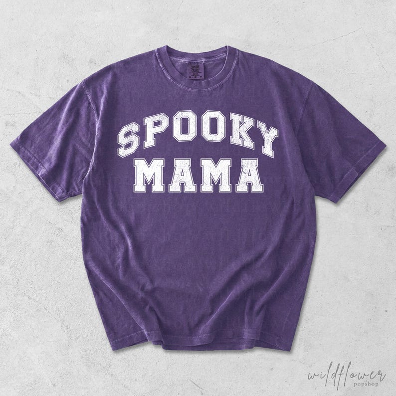May include: A purple short-sleeve t-shirt with a crew neckline. The front features the text SPOOKY MAMA printed in a white, distressed, collegiate-style font. The fabric has a washed, vintage appearance with short sleeves and a standard hem.