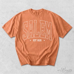 May include: A burnt orange short-sleeve t-shirt featuring the word SALEM in large white outlined capital letters across the chest. Below the text, the words EST 1626 are centered between two small white stars. The shirt has a crew neckline.