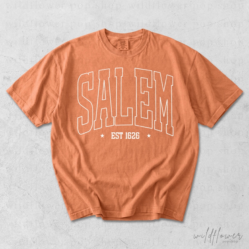 May include: A burnt orange short-sleeve t-shirt featuring the word SALEM in large white outlined capital letters across the chest. Below the text, the words EST 1626 are centered between two small white stars. The shirt has a crew neckline.