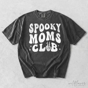 May include: A charcoal gray short-sleeve t-shirt featuring the white distressed text SPOOKY MOMS CLUB printed on the front. The letter U in CLUB is replaced by a white graphic of a skeleton hand making a peace sign, accented by small stars.