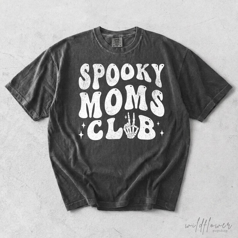 May include: A charcoal gray short-sleeve t-shirt featuring the white distressed text SPOOKY MOMS CLUB printed on the front. The letter U in CLUB is replaced by a white graphic of a skeleton hand making a peace sign, accented by small stars.