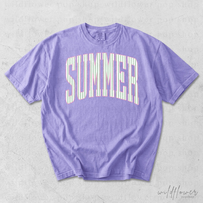 May include: A lilac-colored t-shirt with the word "SUMMER" printed across the front in a white and green striped design. The shirt has short sleeves and a crew neck.