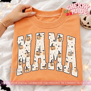 May include: An orange crewneck sweatshirt featuring the word MAMA in large block letters. The letters have a cream-coloured fill patterned with small black stars and orange jack-o'-lanterns wearing black witch hats with a distressed texture.