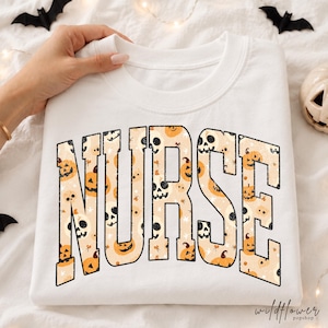 May include: A white crewneck sweatshirt featuring the word NURSE in large, bold, block letters. The letters are filled with a repeating pattern of orange jack-o'-lanterns, white skulls, and small black stars on a light beige background.
