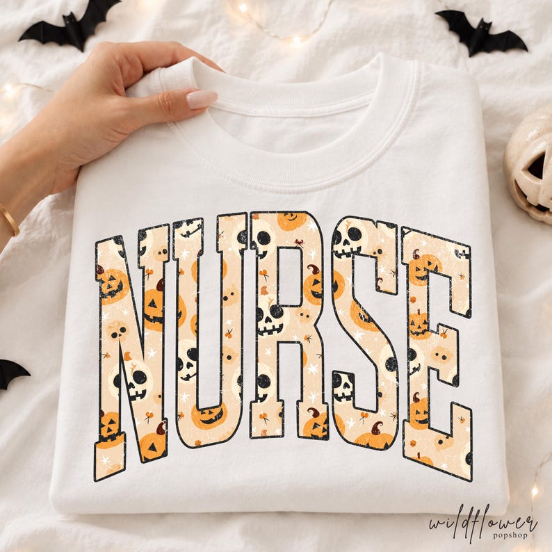 May include: A white crewneck sweatshirt featuring the word NURSE in large, bold, block letters. The letters are filled with a repeating pattern of orange jack-o'-lanterns, white skulls, and small black stars on a light beige background.