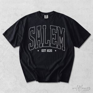 May include: A black short-sleeve t-shirt featuring the word SALEM in large white outlined collegiate-style lettering across the chest. Below the text, the phrase EST 1626 is centered between two small white stars. The shirt has a crew neckline.