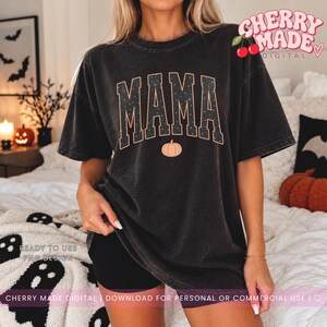 K&ouml;nnte beinhalten: Ein anthrazitfarbenes Kurzarm-T-Shirt mit dem Wort MAMA in gro&szlig;en Blockbuchstaben und einem dezenten Sternenmuster. Unter dem Text befindet sich eine kleine orangefarbene K&uuml;rbisgrafik. Das T-Shirt hat eine lockere Passform und ein Vintage-Finish.
