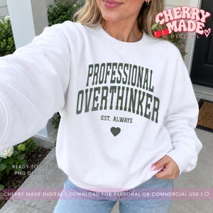 May include: A white crewneck sweatshirt featuring dark green collegiate-style text that reads PROFESSIONAL OVERTHINKER, with the smaller text EST. ALWAYS below it and a small dark green heart icon centered at the bottom of the graphic.