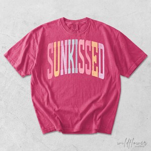 May include: A short-sleeve crew neck t-shirt in a vibrant hot pink color. The front features the word SUNKISSED printed in a large, arched, distressed font with alternating pastel colors including yellow, light pink, and light blue.