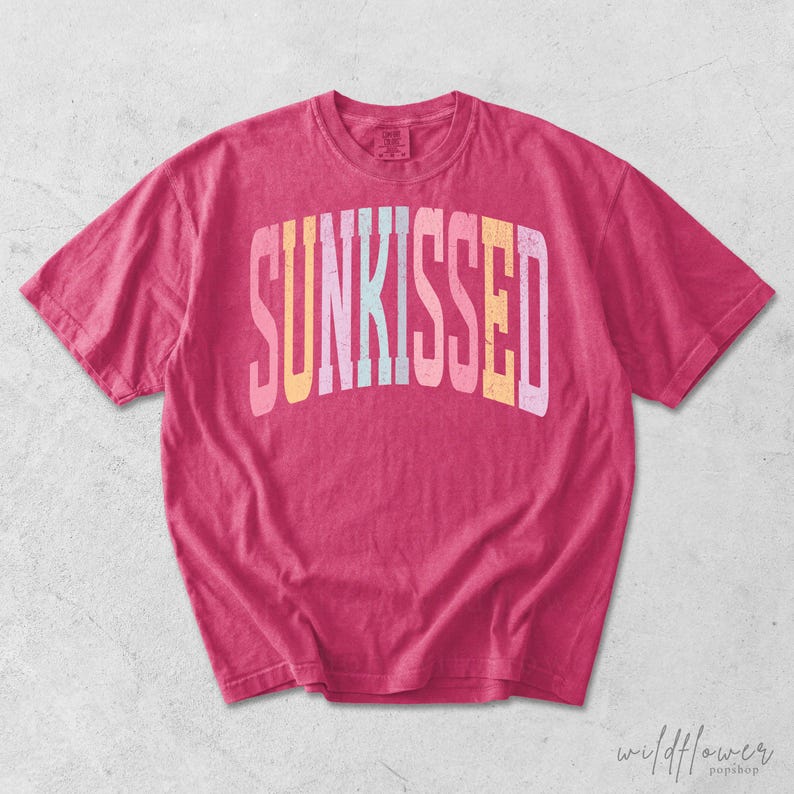 May include: A short-sleeve crew neck t-shirt in a vibrant hot pink color. The front features the word SUNKISSED printed in a large, arched, distressed font with alternating pastel colors including yellow, light pink, and light blue.