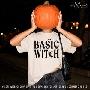 May include: A cream-colored short-sleeve t-shirt featuring the black text BASIC WITCH with a small bat and crescent moon graphic. The shirt is paired with dark denim jeans and a black shoulder bag, while a large orange pumpkin is held up.