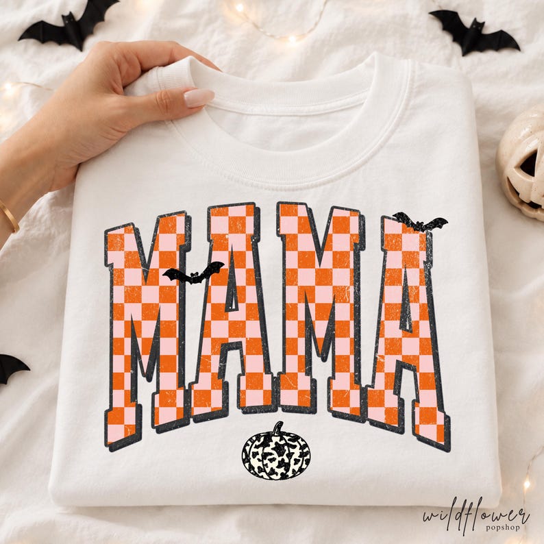 Puede incluir: Una sudadera blanca de cuello redondo con la palabra MAMA en un patr&oacute;n de cuadros naranja y rosa claro desgastado. Las letras est&aacute;n delineadas en negro, con dos peque&ntilde;os murci&eacute;lagos negros sobre las letras y una calabaza blanca y negra.