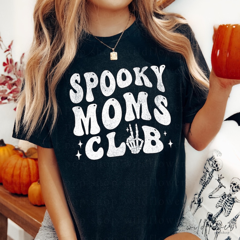 May include: A black short-sleeve t-shirt featuring white distressed text that reads SPOOKY MOMS CLUB, with a skeleton hand making a peace sign replacing the letter U in CLUB, accented by small four-pointed star graphics.