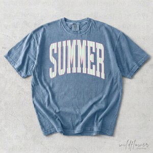 May include: A blue t-shirt with the word "SUMMER" in large, white, striped letters across the front. The stripes are light green and pink. The shirt has a crew neck and short sleeves.