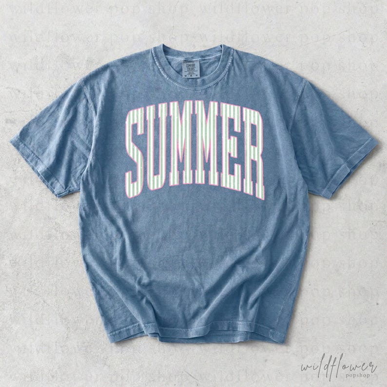 May include: A blue t-shirt with the word "SUMMER" in large, white, striped letters across the front. The stripes are light green and pink. The shirt has a crew neck and short sleeves.