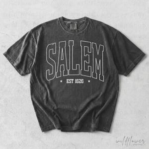 May include: A charcoal gray short-sleeve t-shirt with a crew neckline. The front features white outlined block lettering that reads SALEM, with the text EST 1626 centered below it, flanked by two small five-pointed stars on either side.