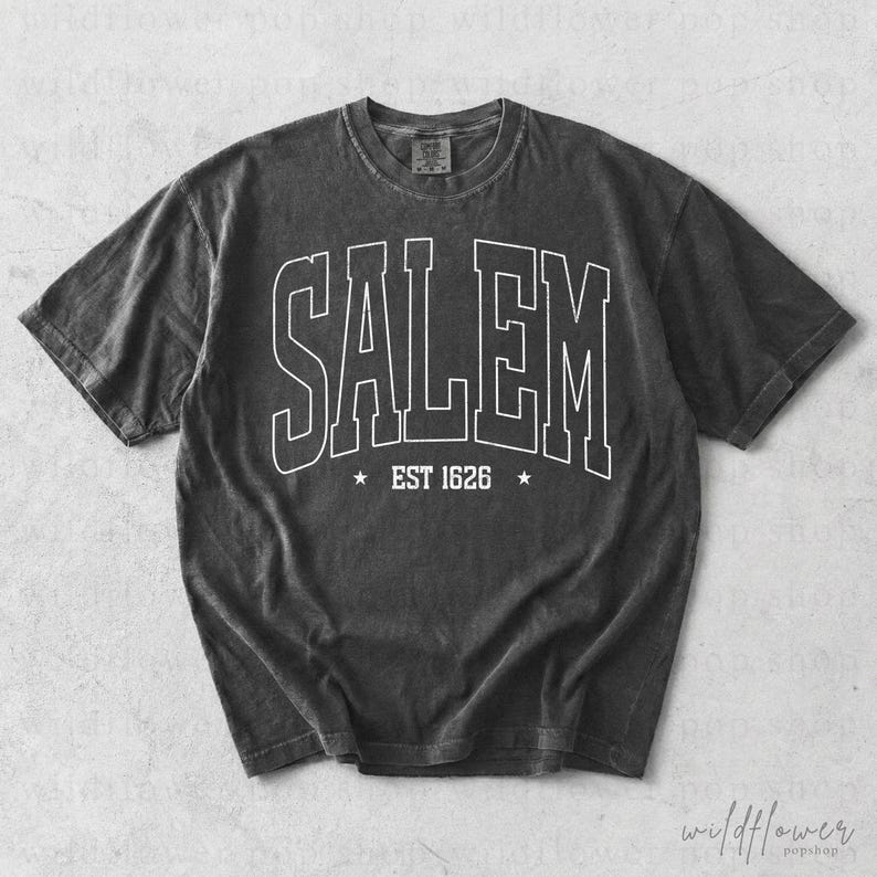 May include: A charcoal gray short-sleeve t-shirt with a crew neckline. The front features white outlined block lettering that reads SALEM, with the text EST 1626 centered below it, flanked by two small five-pointed stars on either side.